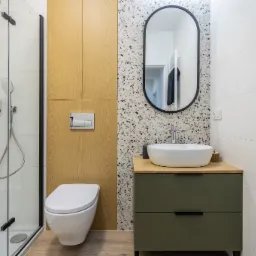 Nowoczesna łazienka z kabiną prysznicową, toaletą wiszącą, umywalką nablatową na szafce z dwiema szufladami w kolorze oliwkowym, lustrem owalnym w czarnej ramie oraz ścianą w mozaikę lastryko.