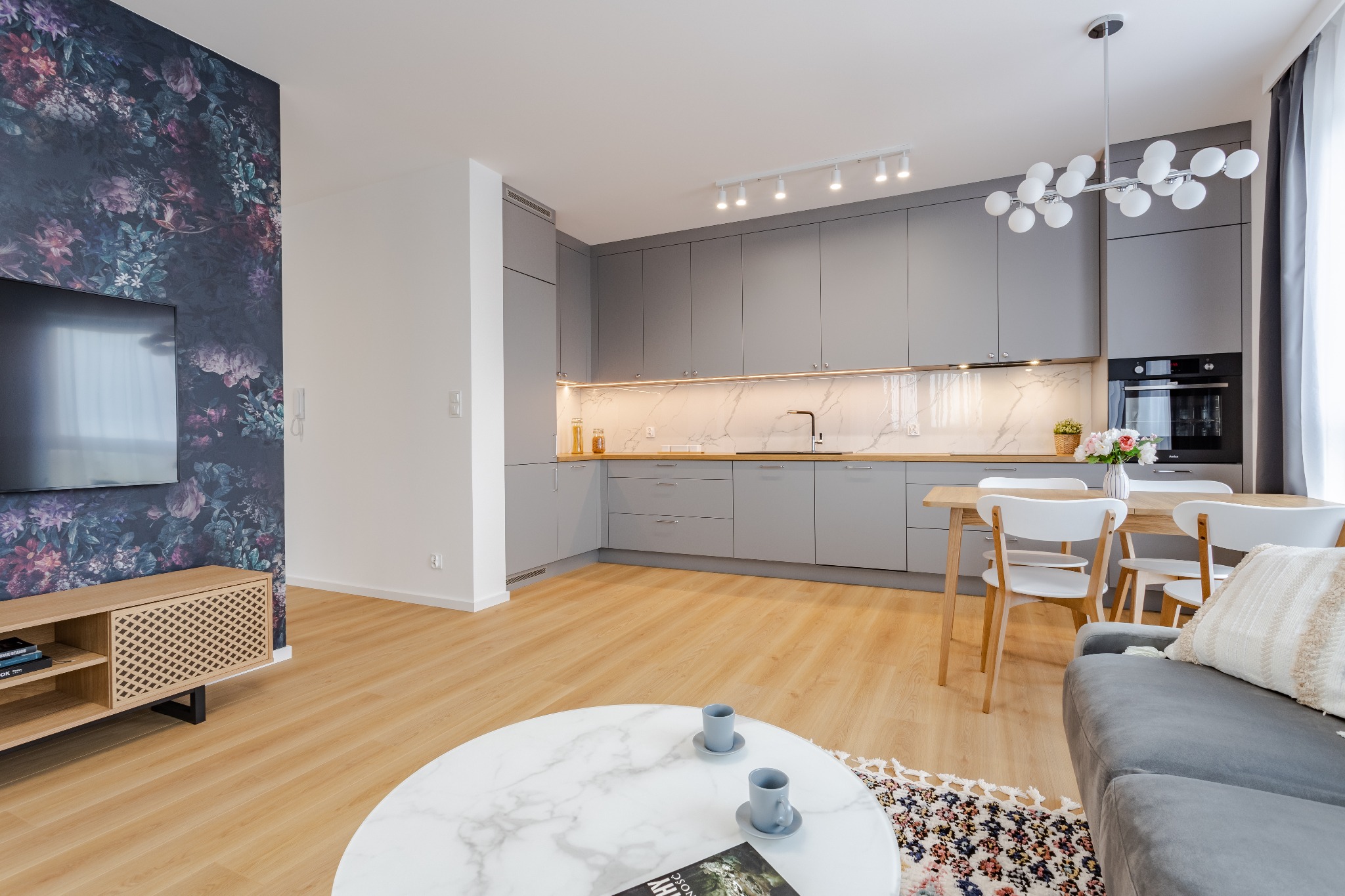 Nowoczesna szara kuchnia z drewnianym blatem i marmurowym backsplashiem, widok na jadalnię z drewnianym stołem i białymi krzesłami, okrągły stolik kawowy z marmurowym blatem i dwoma filiżankami...