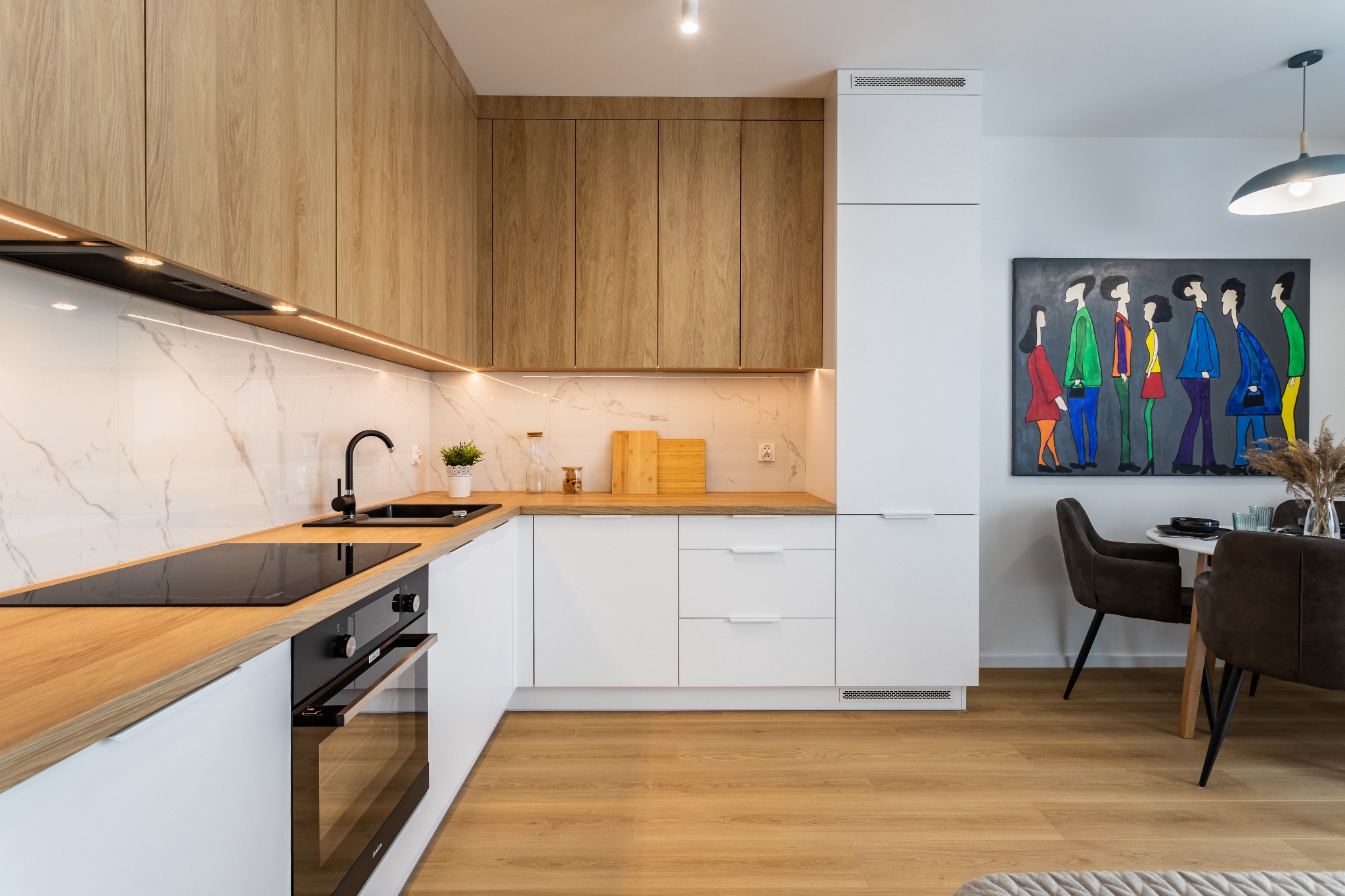 Nowoczesna kuchnia w jasnych barwach: białe szafki dolne z drewnianym blatem, górne szafki z drewnianymi frontami, marmurowe płytki na ścianie, czarna armatura i płyta indukcyjna, stół z krzesłami...