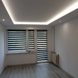 Pusty pokój po remoncie z panelami podłogowymi w odcieniach szarości, białymi ścianami, oknem z żaluzjami dzień-noc i podwieszanym sufitem z oświetleniem LED.