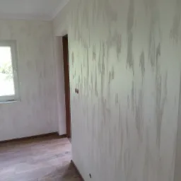 Pomieszczenie po malowaniu ścian z efektem dekoracyjnym w odcieniach szarości, widoczna drewnopodobna podłoga, okno i fragment futryny drzwiowej.