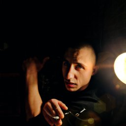 GMG - Portret młodego mężczyzny z krótkimi włosami w ciemnym otoczeniu, oświetlonego bocznym światłem lampy, trzymającego w dłoni zapalonego papierosa, z widocznym dymem.