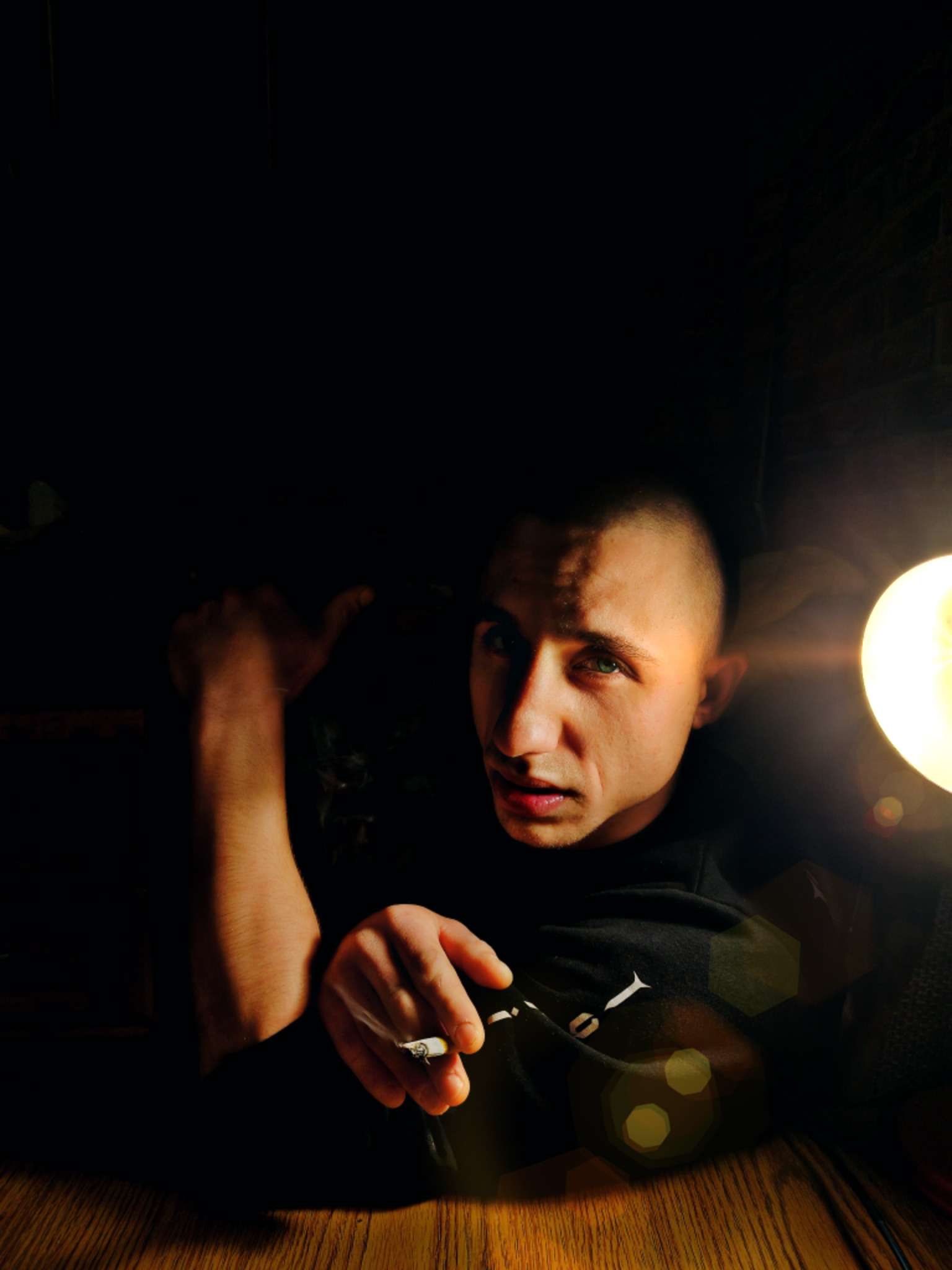 Portret młodego mężczyzny z krótkimi włosami w ciemnym otoczeniu, oświetlonego bocznym światłem lampy, trzymającego w dłoni zapalonego papierosa, z widocznym dymem.