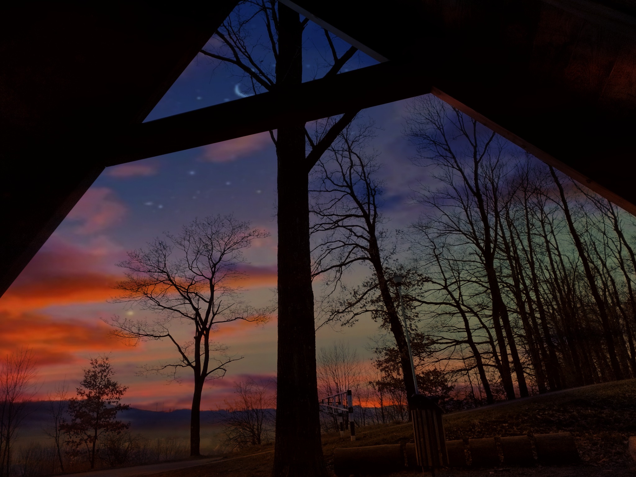 Widok na krajobraz z wnętrza budynku przez drewnianą konstrukcję, zachód słońca z lewej strony, księżyc z prawej, drzewa bez liści na pierwszym planie.