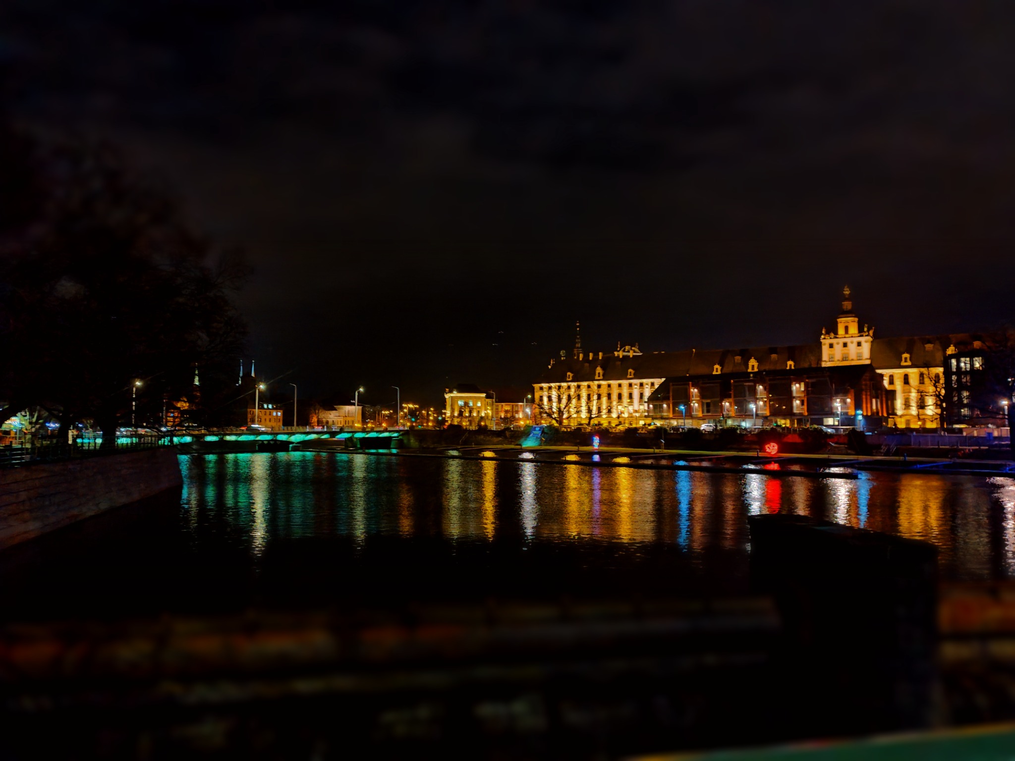 Nocny widok na oświetlony budynek uniwersytetu odbijający się w tafli rzeki, z zielonym oświetleniem mostu w tle.