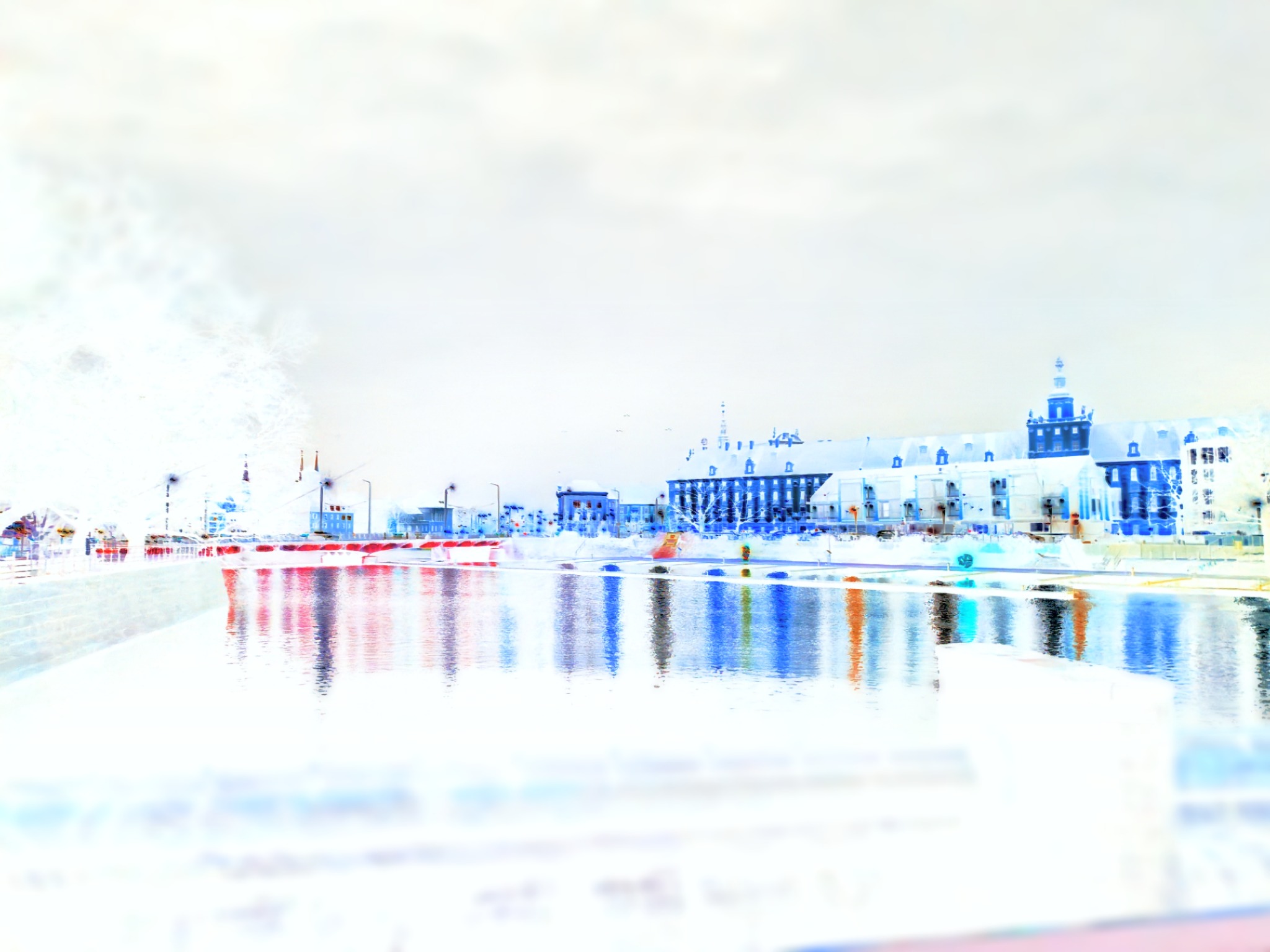 Odwrócone kolorystycznie zdjęcie przedstawia budynki odbijające się w wodzie. W tle widoczny budynek z wieżyczką.