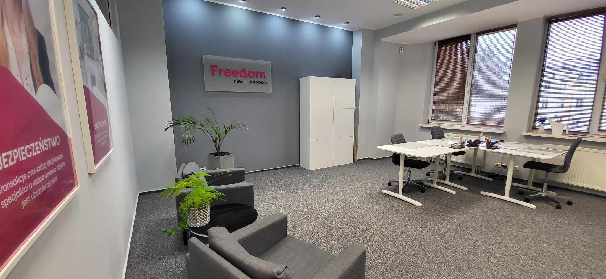 Jasne biuro agencji nieruchomości z szarymi ścianami, biurkami, krzesłami i roślinami doniczkowymi; logo 'Freedom nieruchomości' na ścianie.