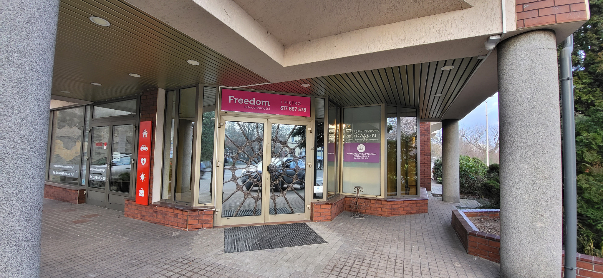 Wejście do biura nieruchomości Freedom z dekoracyjnymi, metalowymi zdobieniami na szklanych drzwiach, widoczny szyld z numerem telefonu i logo firmy.