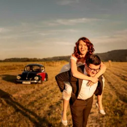 Uśmiechnięta panna młoda na barana u pana młodego na polu z klasycznym samochodem VW Garbus w tle, w promieniach zachodzącego słońca.