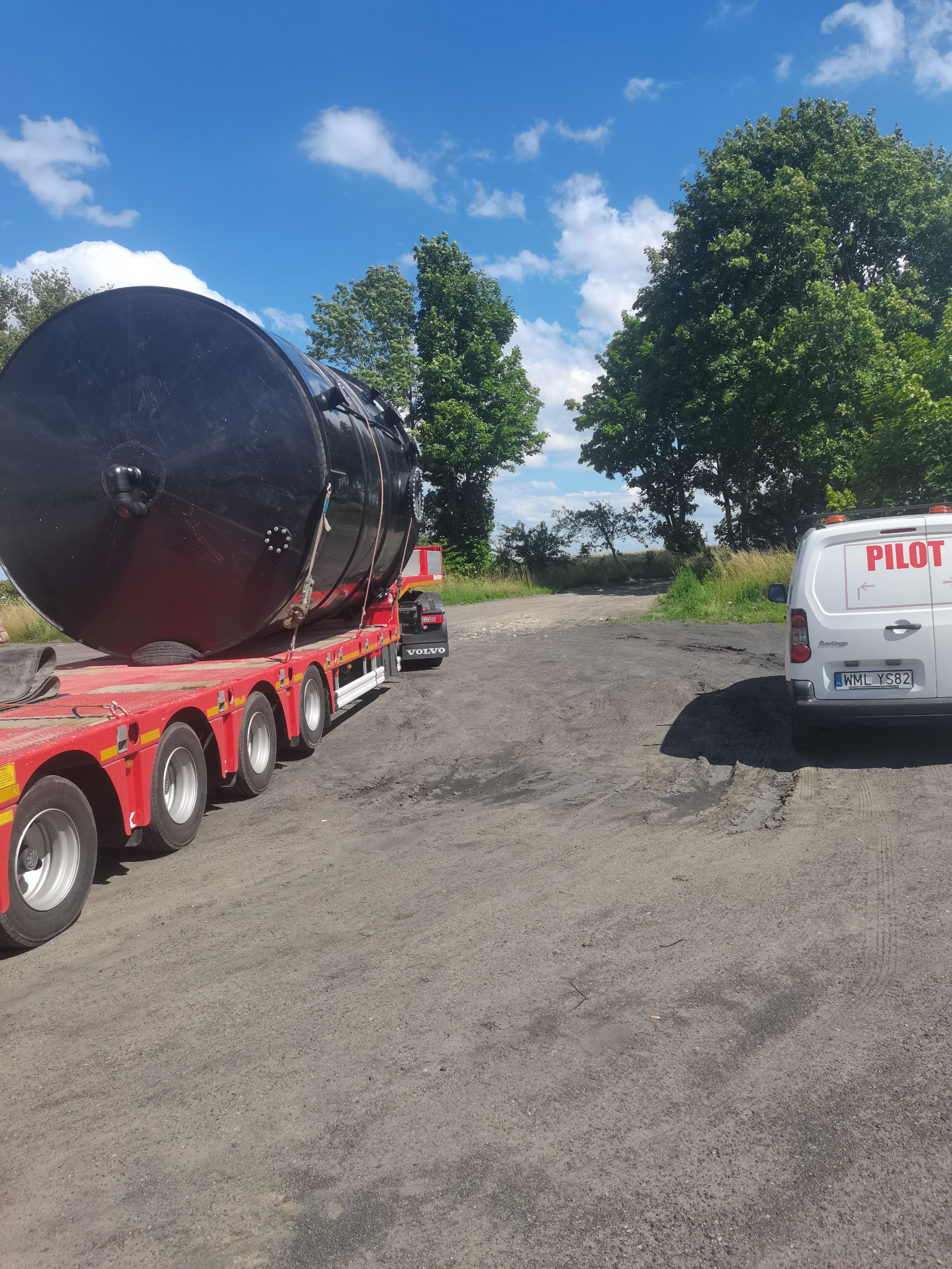 Czarny zbiornik przemysłowy zabezpieczony na czerwonej platformie niskopodwoziowej Volvo, widoczny samochód pilotujący z napisem PILOT, droga gruntowa, błękitne niebo z chmurami.