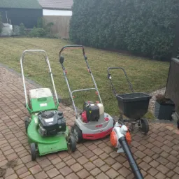 Dwie kosiarki spalinowe, siewnik do trawy i dmuchawa do liści stoją na kostce brukowej przed trawnikiem i żywopłotem.