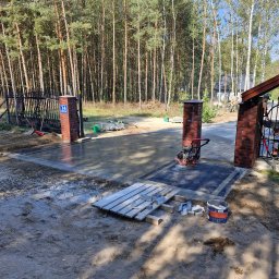 Nie Podano - Wjazd do posesji z częściowo ułożoną, szarą kostką brukową, zagęszczarką wibro płytową, ceglanymi słupkami i lasem w tle, widoczne narzędzia i materiały budowlane.