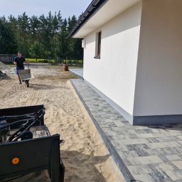 Nie Podano - Mężczyzna z taczką na piasku w trakcie układania szarej kostki brukowej przy białym budynku z ciemnym dachem i rynną.