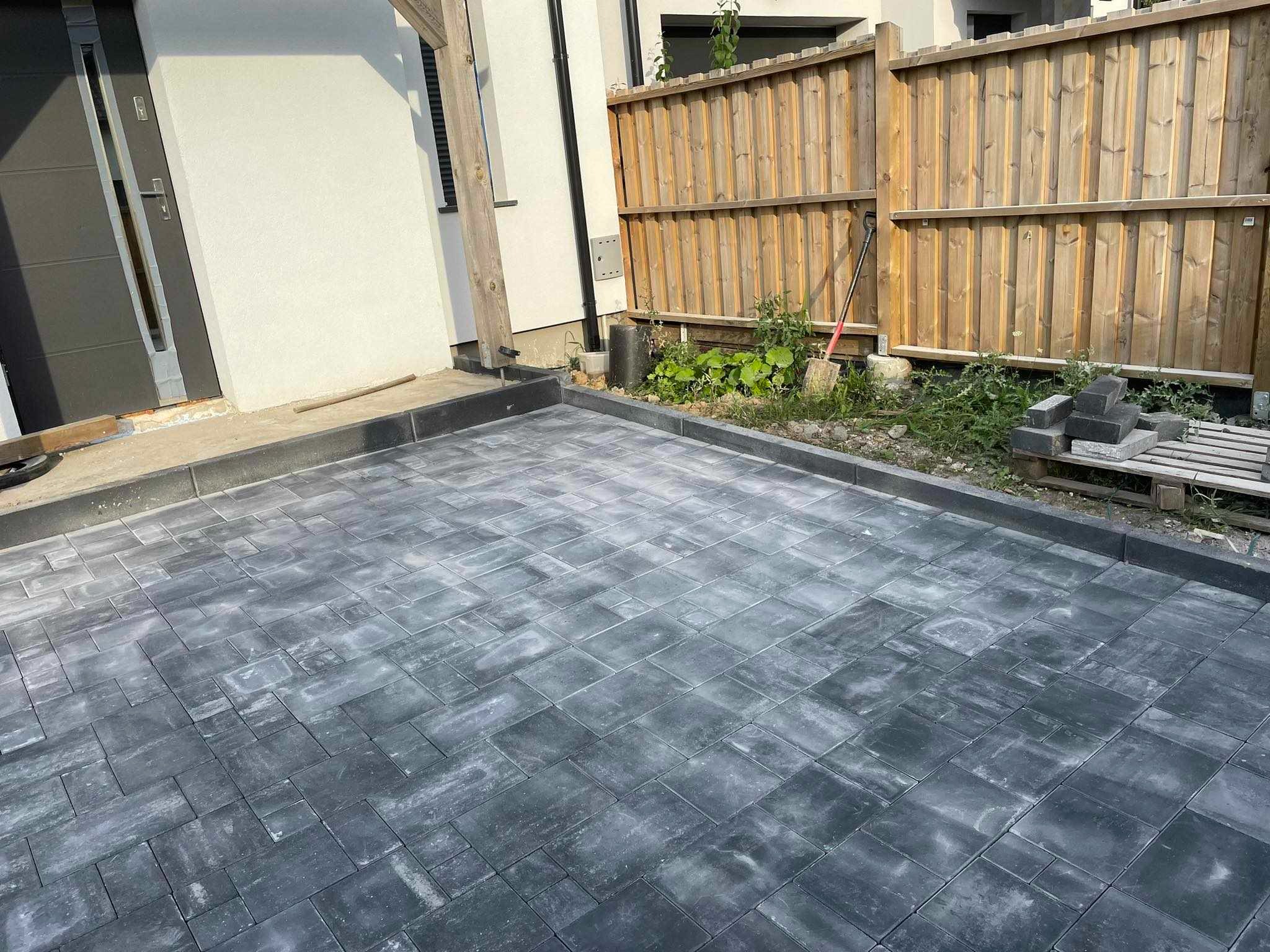 Ułożona szara kostka brukowa na podjeździe, widoczna drewniana brama, dom z szarą stolarką okienną i drzwiową, w tle zielona roślinność.