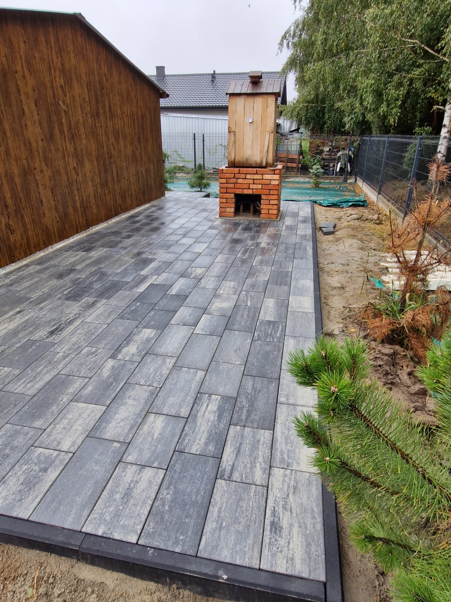 Ułożona szara kostka brukowa z czarnym obrzeżem, obok drewniana wędzarnia z ceglaną podstawą, widok z poziomu gruntu.