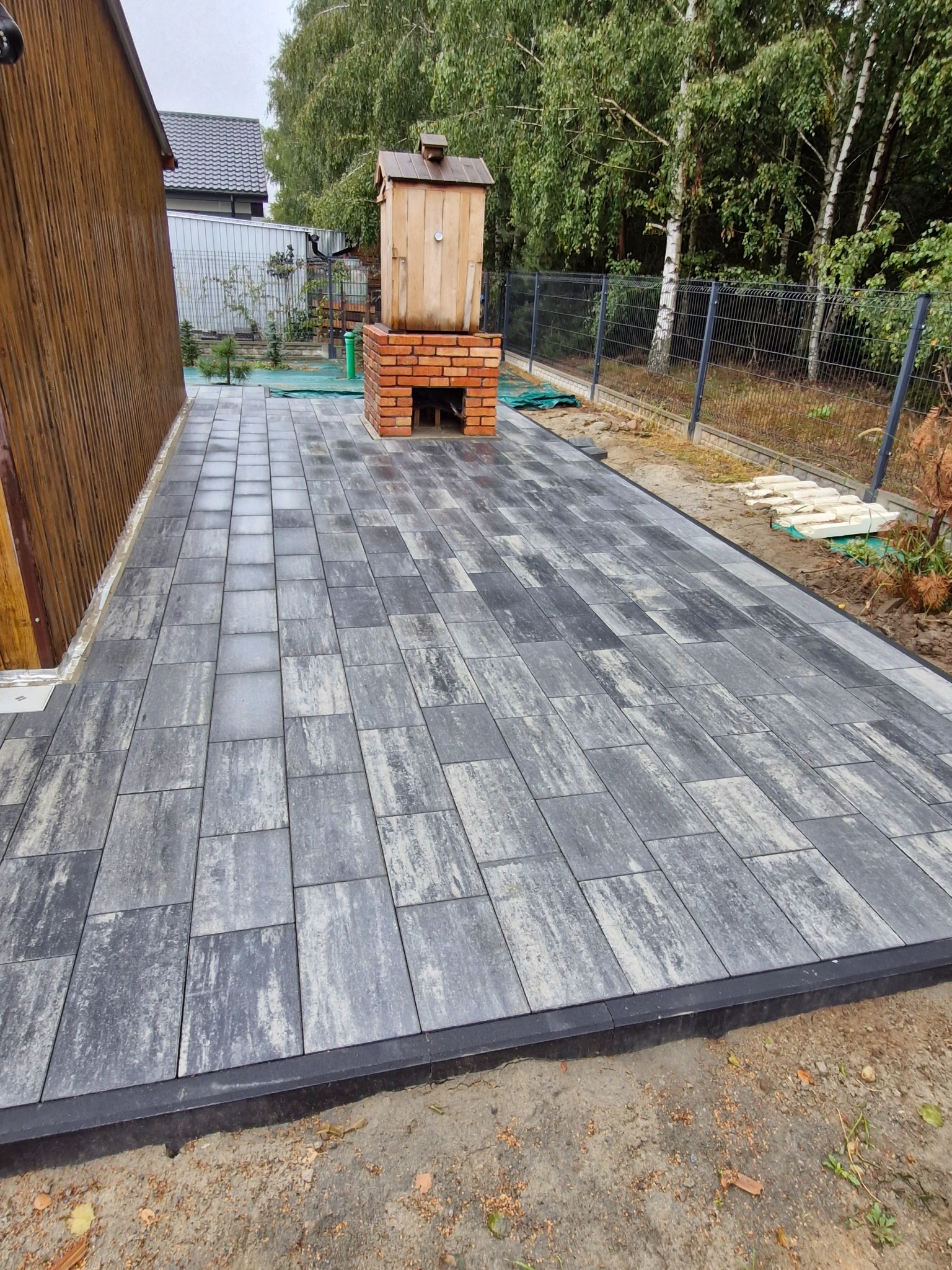 Ułożona szara kostka brukowa z widoczną wędzarnią z cegły i drewna w tle, obrzeża z czarnego tworzywa sztucznego, teren jeszcze niezagospodarowany.
