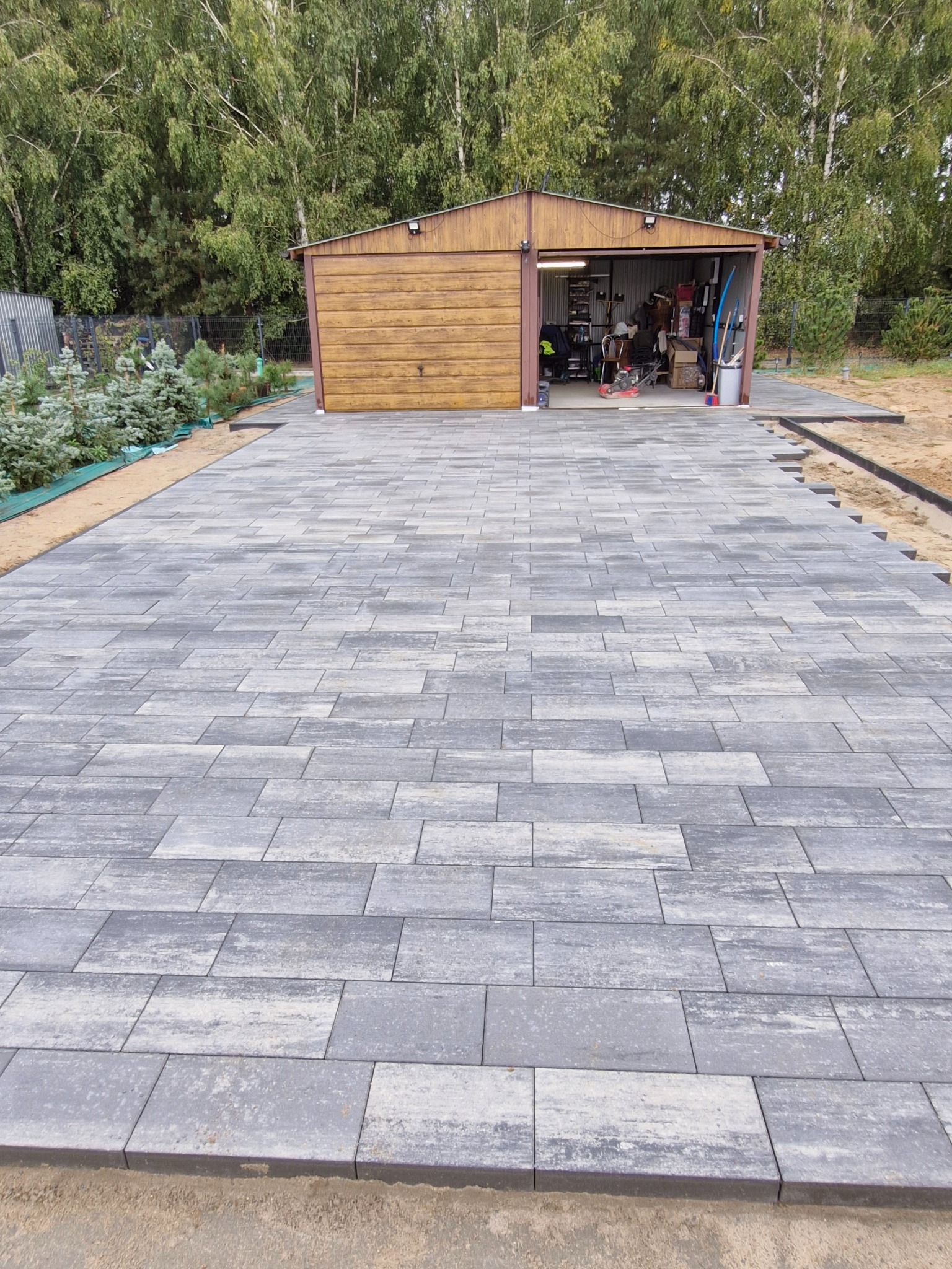Ułożona szara kostka brukowa prowadząca do garażu z drewnianą bramą, widoczne obrzeża i przygotowane podłoże.