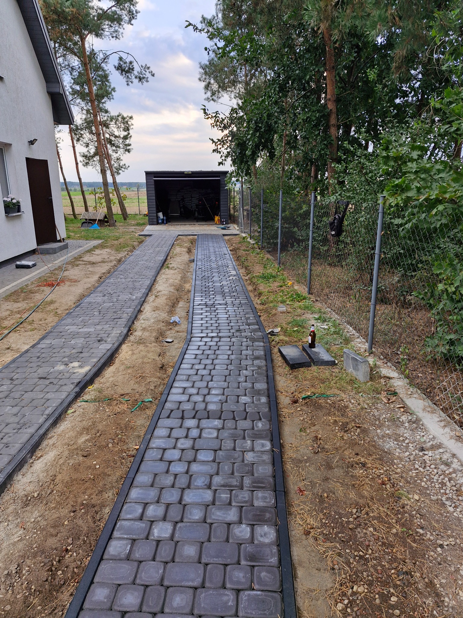 Dwa równoległe chodniki z szarej kostki brukowej prowadzące do garażu, z widoczną butelką piwa obok ogrodzenia.