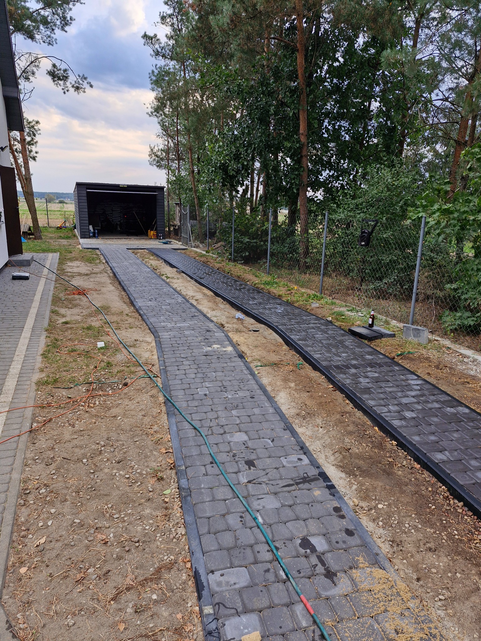 Dwie ścieżki z szarej kostki brukowej prowadzące do garażu, jedna ułożona, druga w trakcie układania, widoczne narzędzia i butelka po piwie przy ogrodzeniu.