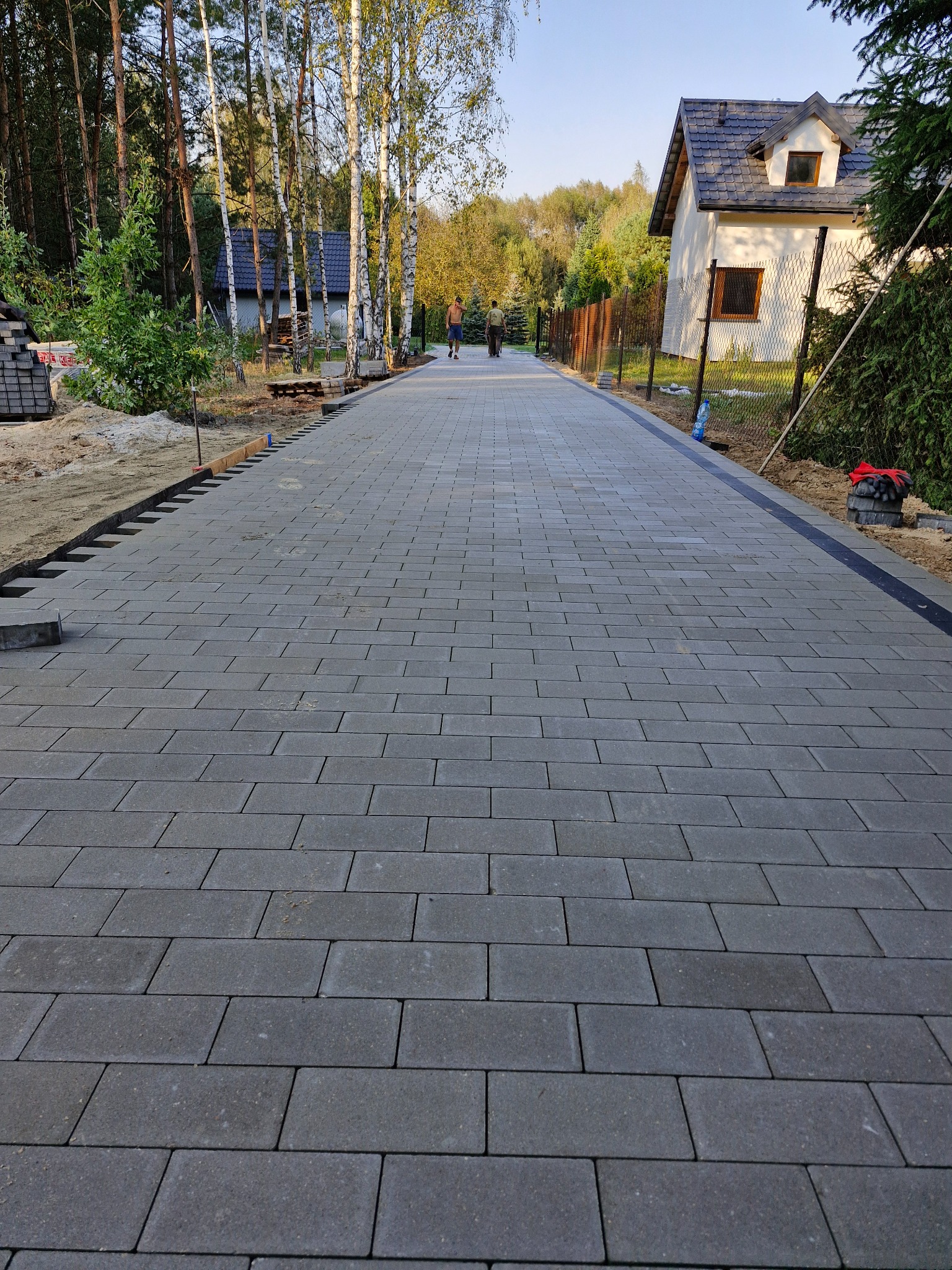 Długa, szara ścieżka z kostki brukowej prowadząca przez posesję z domem i drzewami, widoczne przygotowania do dalszego układania kostki po lewej stronie.