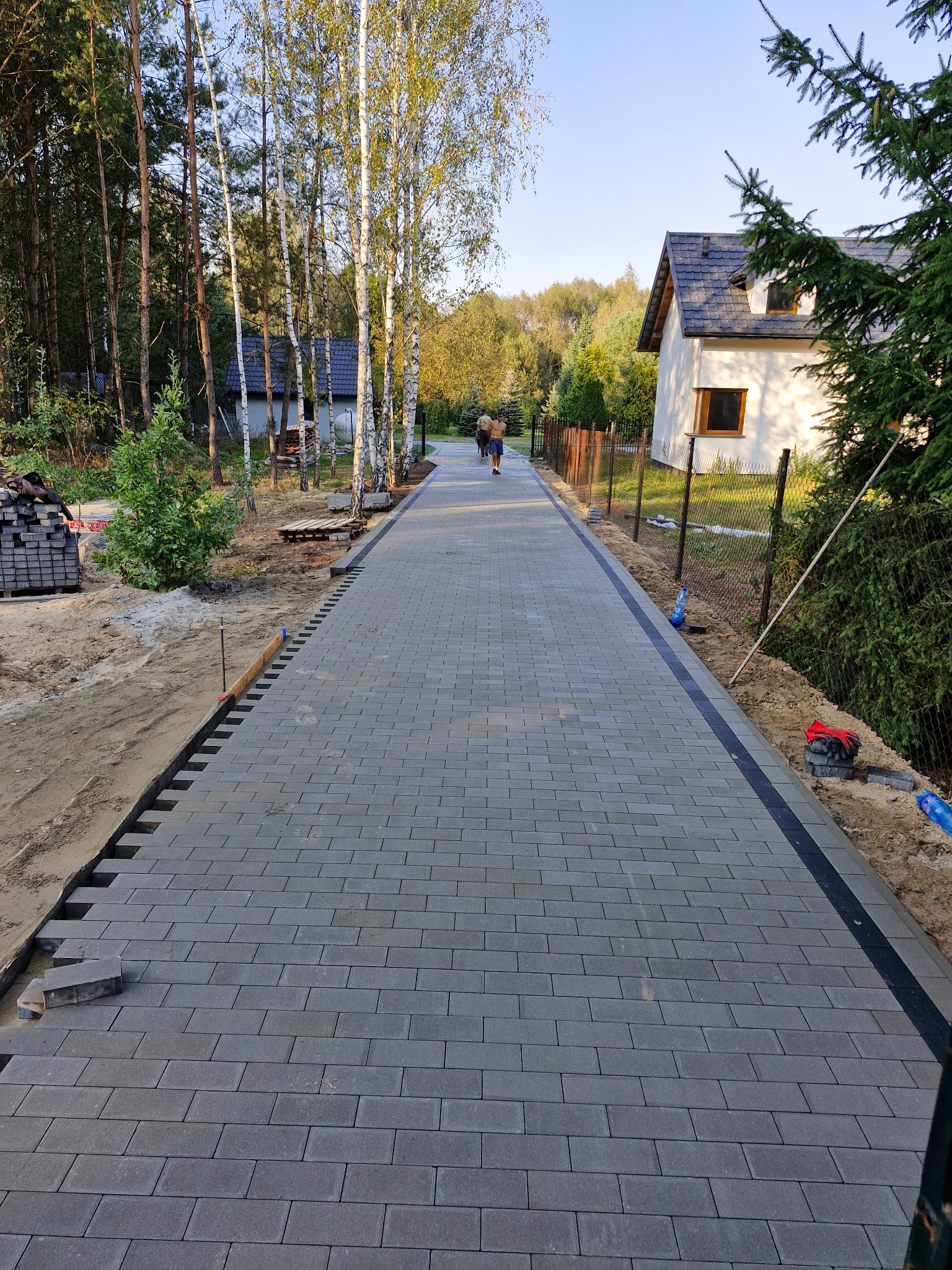Długa, szara ścieżka z kostki brukowej prowadząca przez posesję z brzozami i domem w tle, widoczne niedokończone obrzeża i stos kostki.