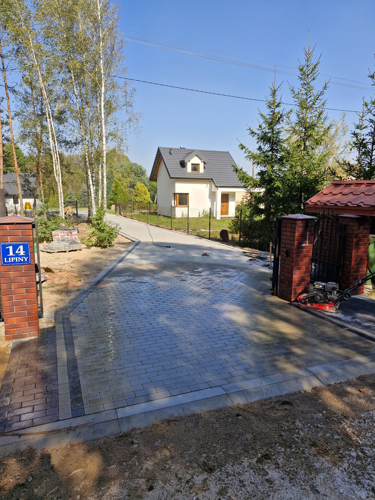 Nowo ułożona, szara kostka brukowa na podjeździe do domu jednorodzinnego, z widocznym numerem posesji '14 Lipiny' na ceglanym słupku bramy, w tle dom z szarym dachem i zielone drzewa.