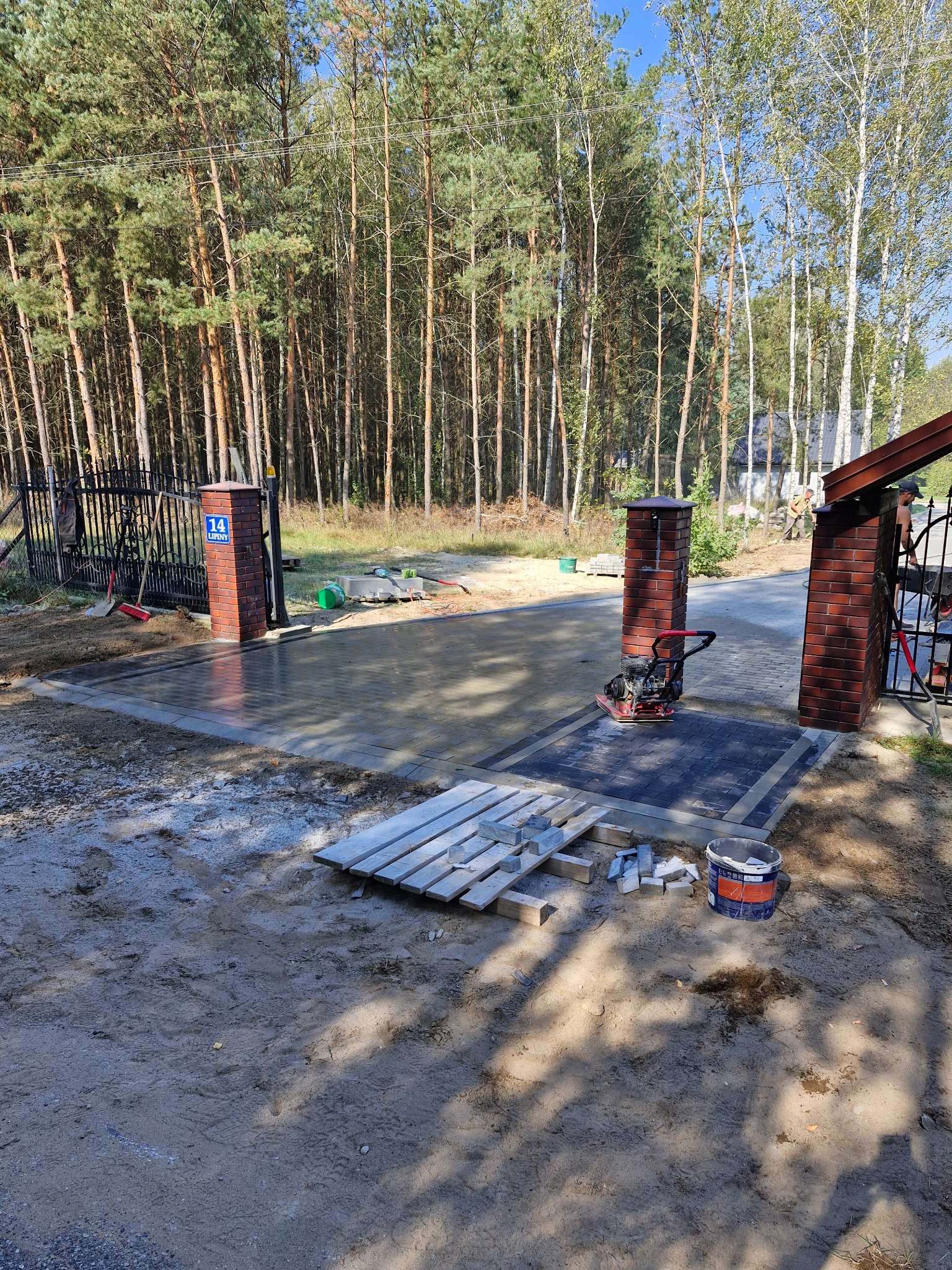 Wjazd do posesji z częściowo ułożoną, szarą kostką brukową, zagęszczarką wibro płytową, ceglanymi słupkami i lasem w tle, widoczne narzędzia i materiały budowlane.