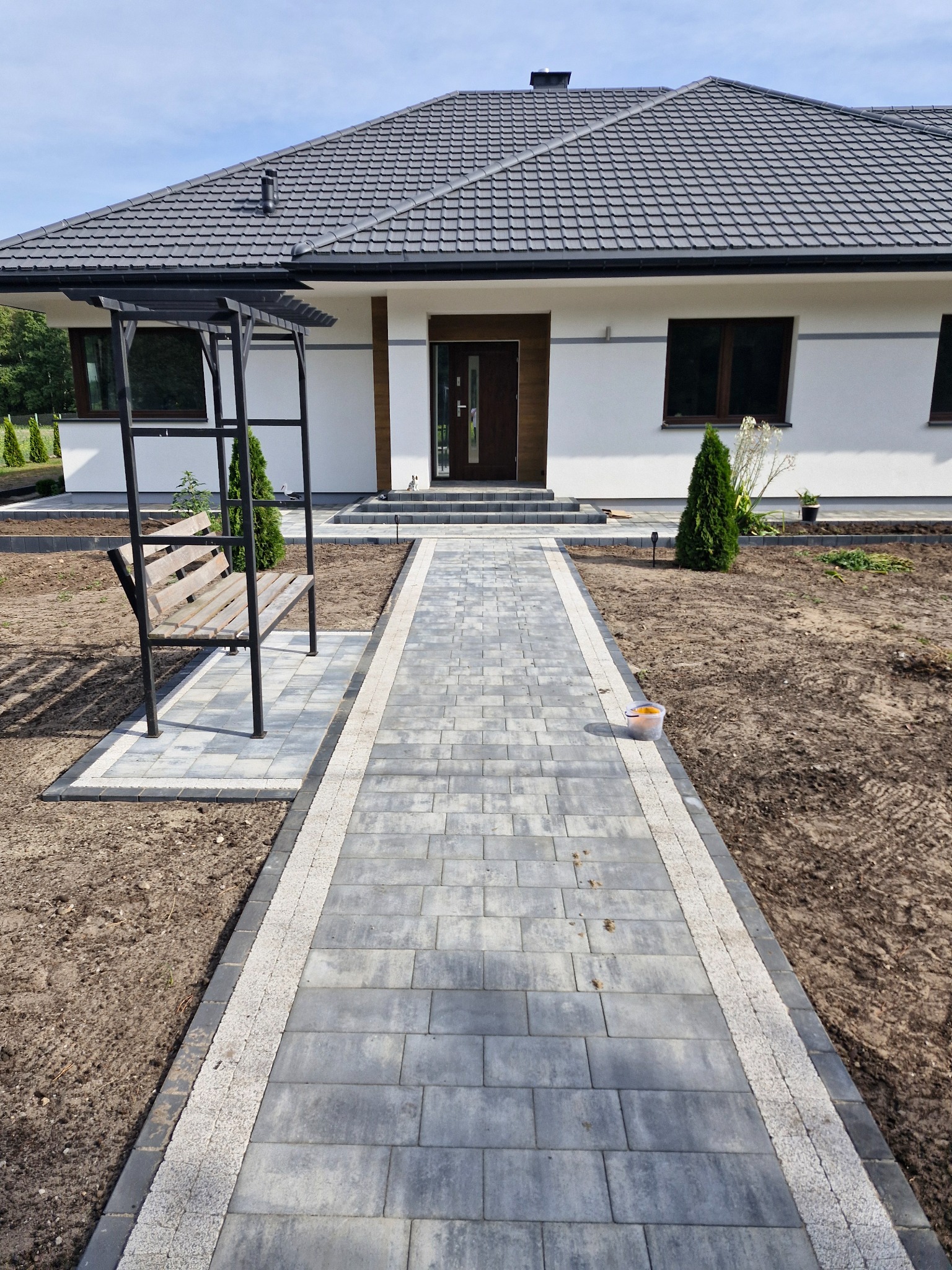 Ułożona kostka brukowa prowadząca do domu z drewnianą pergolą i ławką, widoczne elementy wykończeniowe ogrodu.