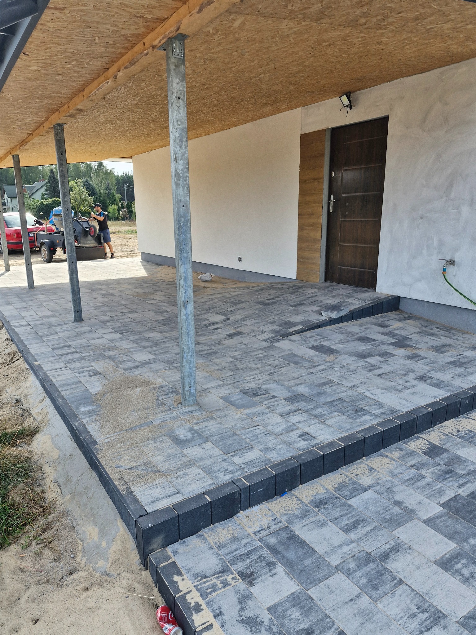Szara kostka brukowa ułożona na podjeździe, widoczne metalowe podpory zadaszenia i pracownik z taczką w tle.