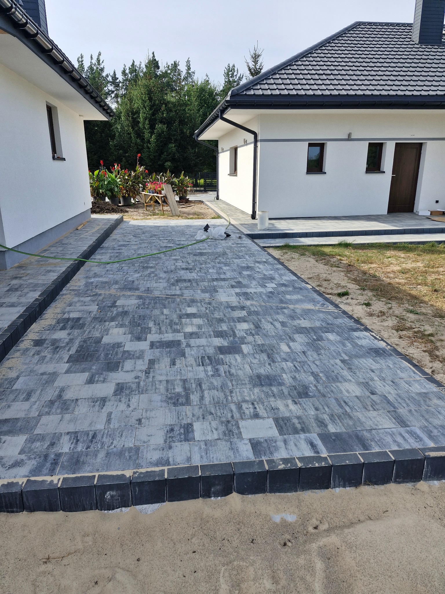 Ułożona szara kostka brukowa na podjeździe, obrzeża z ciemnej kostki, widoczny wąż ogrodowy i biały dom z ciemnym dachem w tle.