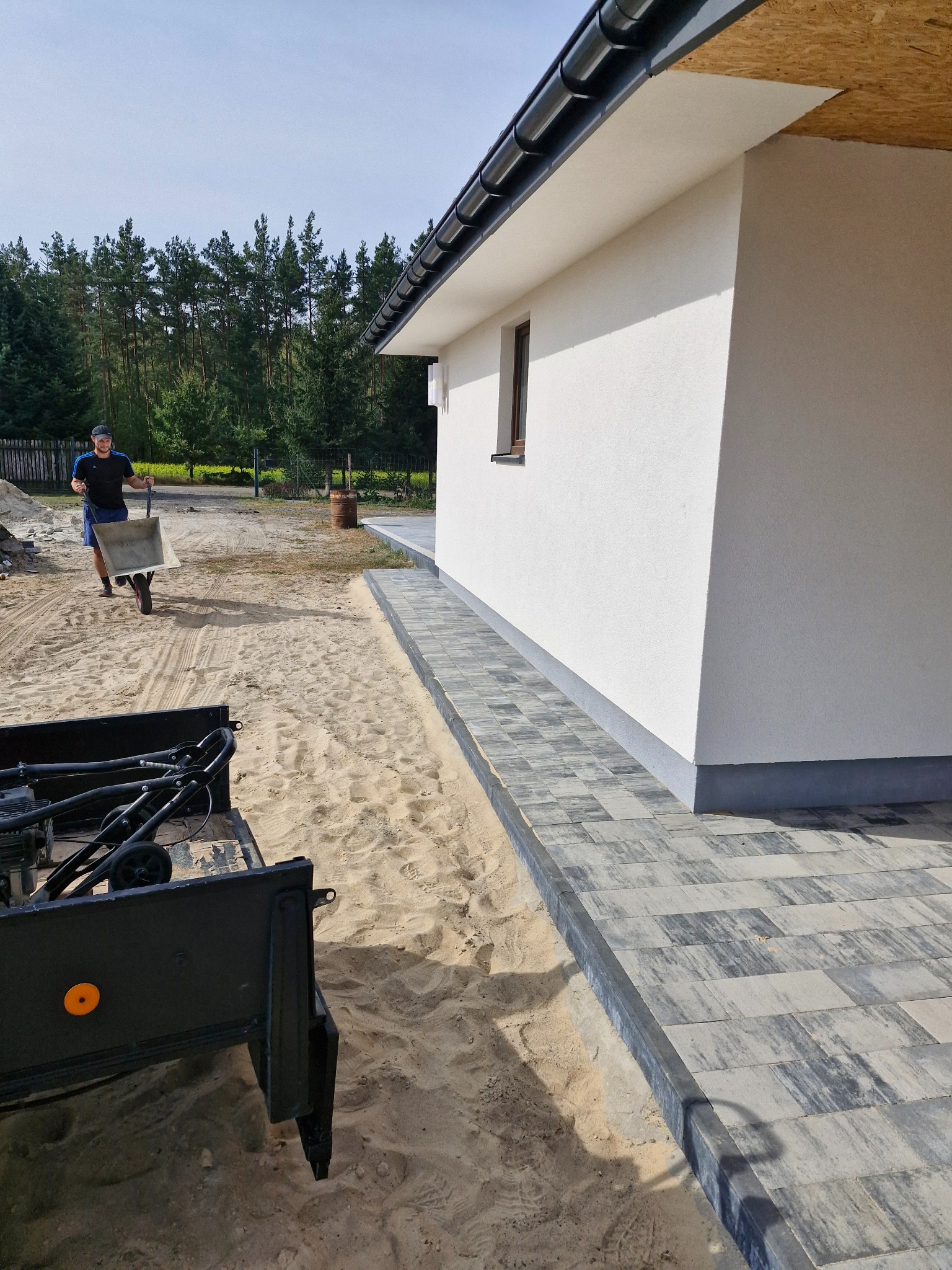 Mężczyzna z taczką na piasku w trakcie układania szarej kostki brukowej przy białym budynku z ciemnym dachem i rynną.