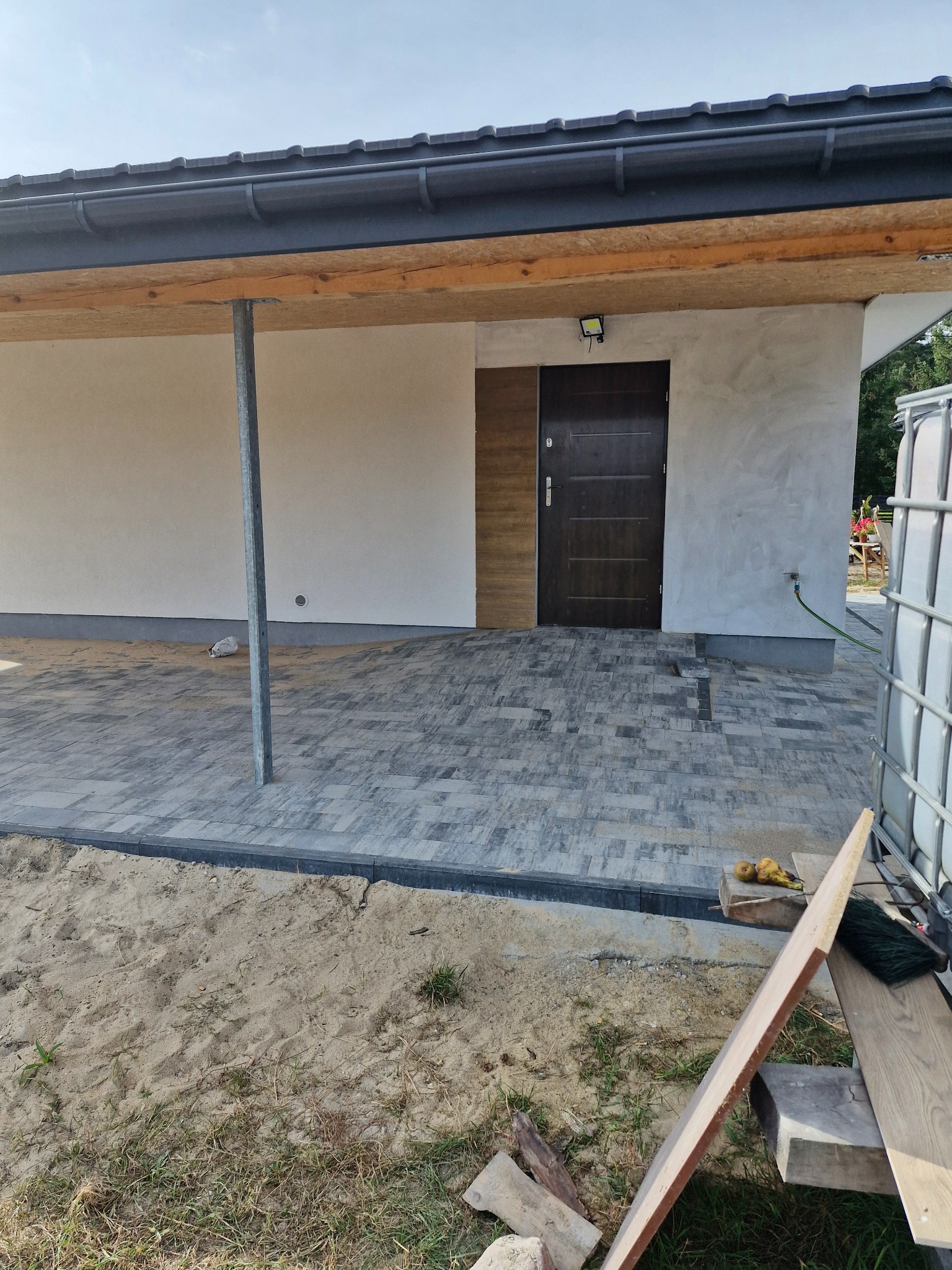 Nowo ułożona szara kostka brukowa przed domem z drewnianymi elementami elewacji, widoczne narzędzia i piasek.