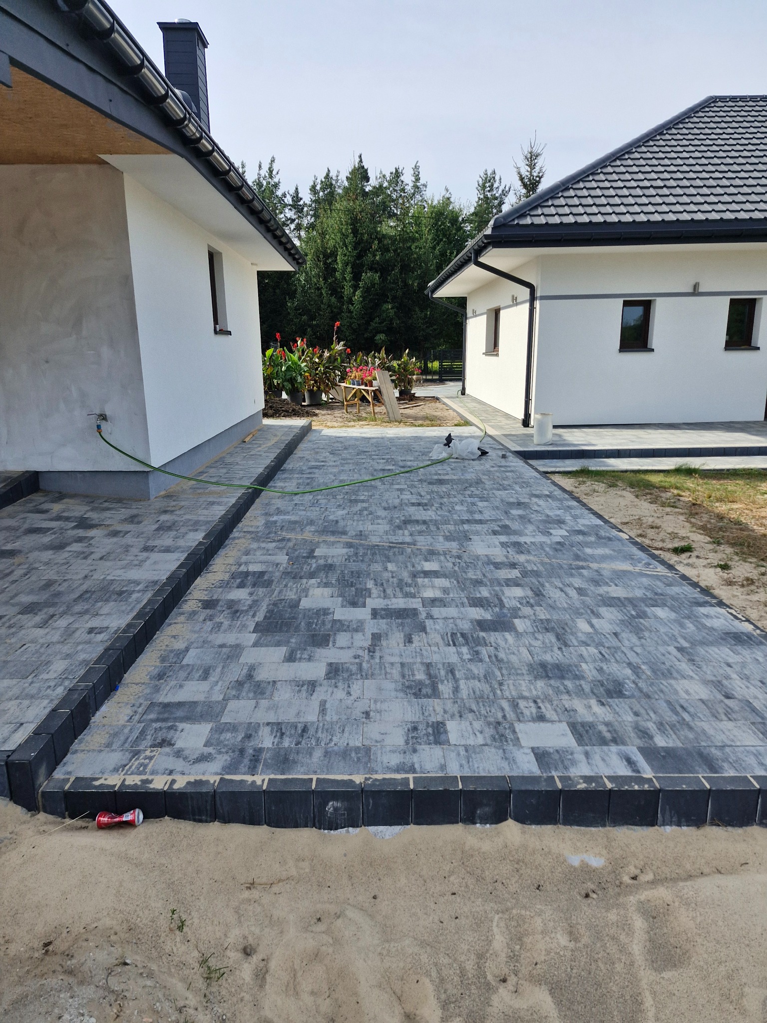 Ułożona szara kostka brukowa prowadząca między dwoma białymi budynkami, widoczny zielony wąż ogrodowy i puszka po napoju na piasku.