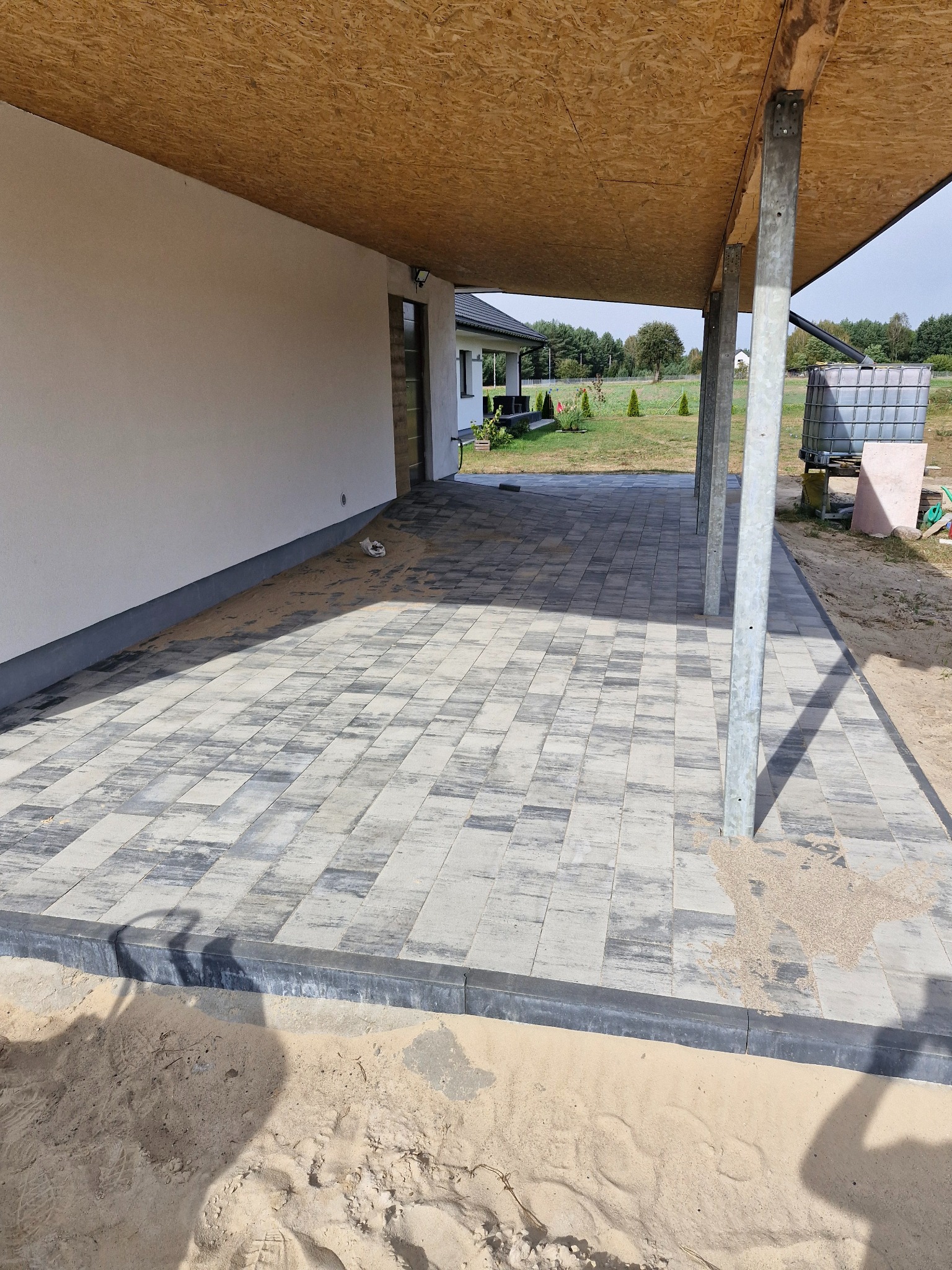 Zadaszony taras z szarej kostki brukowej ułożonej we wzór jodełki, obrzeża z ciemnych kostek, widoczne słupy podtrzymujące zadaszenie i piasek wokół.