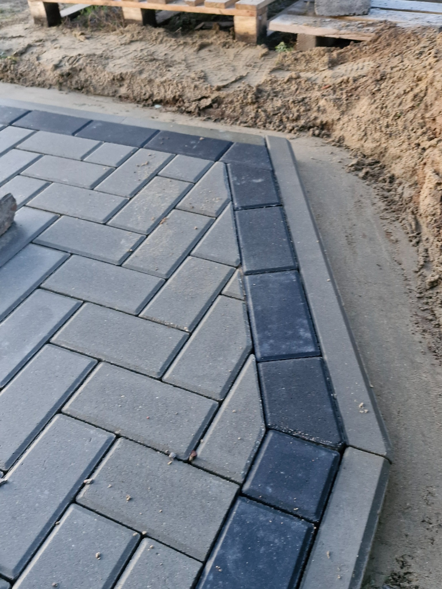 Ułożona kostka brukowa w szarych odcieniach, wzór jodełki, obrzeże z ciemniejszych kostek, widoczny piasek i paleta w tle.