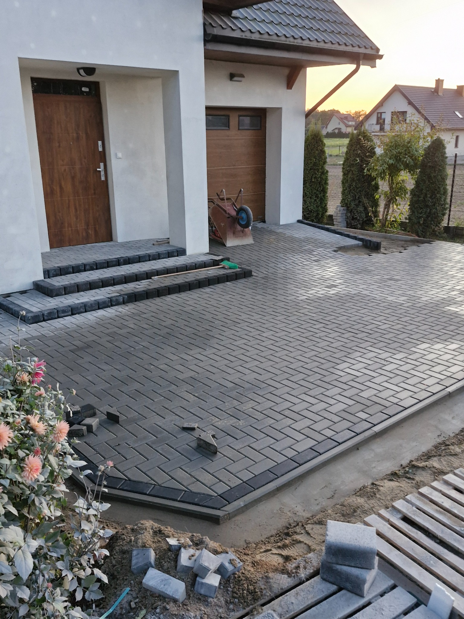 Nowa, szara kostka brukowa ułożona w jodełkę na podjeździe przed domem, widoczne schody wejściowe i częściowo ułożone obrzeża, w tle zachodzące słońce.