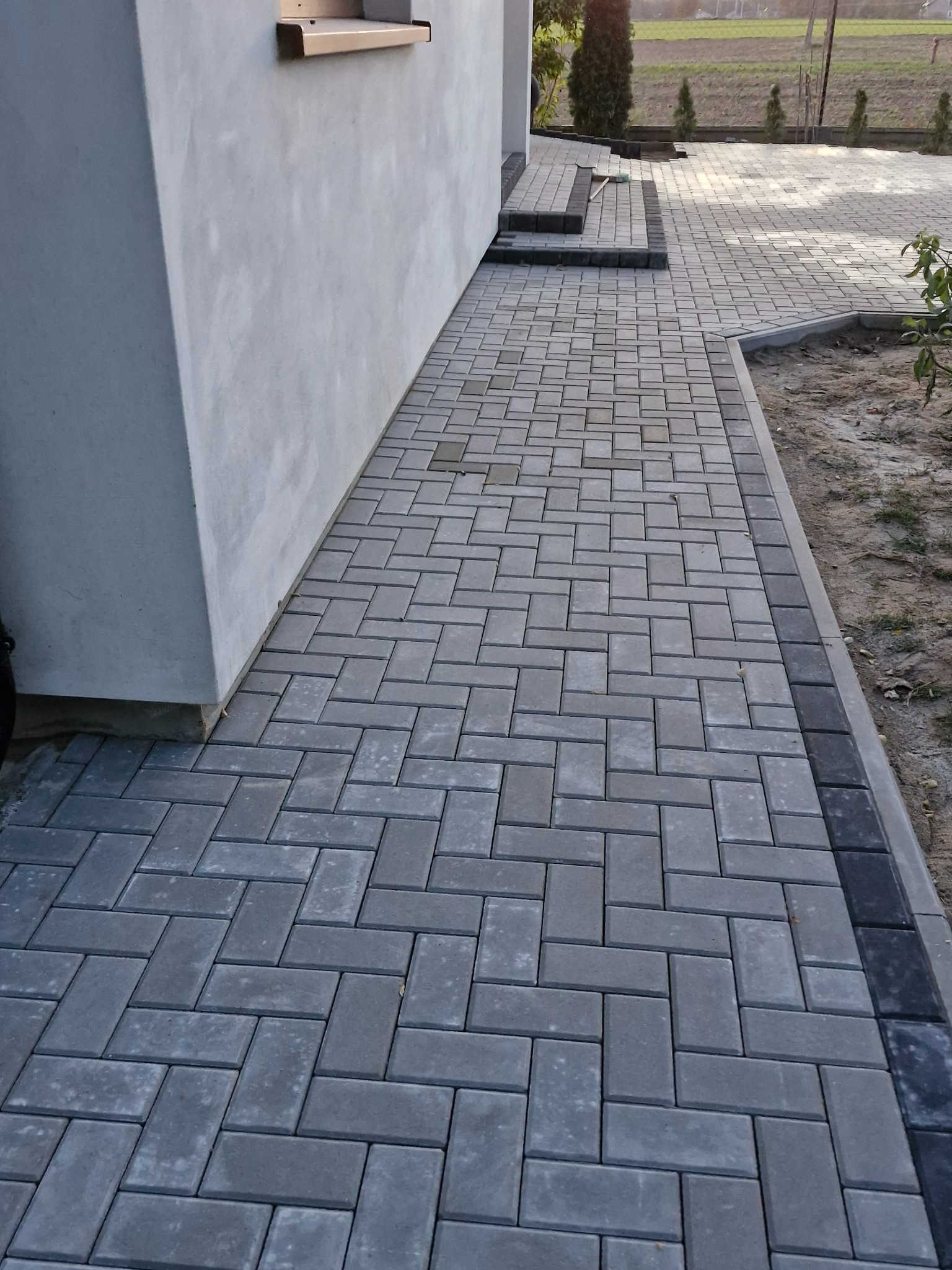 Ułożona szara kostka brukowa w jodełkę tworząca chodnik przy budynku z widocznymi stopniami i obrzeżem z ciemniejszej kostki.