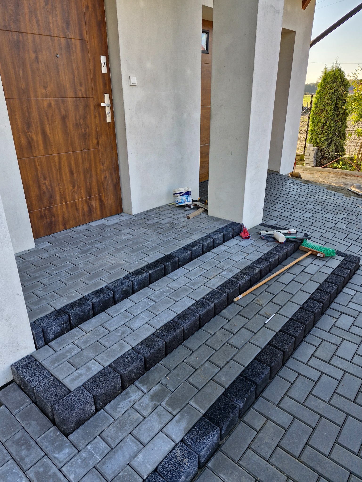 Fragment nowo ułożonej kostki brukowej przed wejściem do domu, z widocznymi narzędziami: poziomica, szczotka, rękawice robocze, puszka farby. Czarna kostka obrzeżna oddziela szarą kostkę główną.