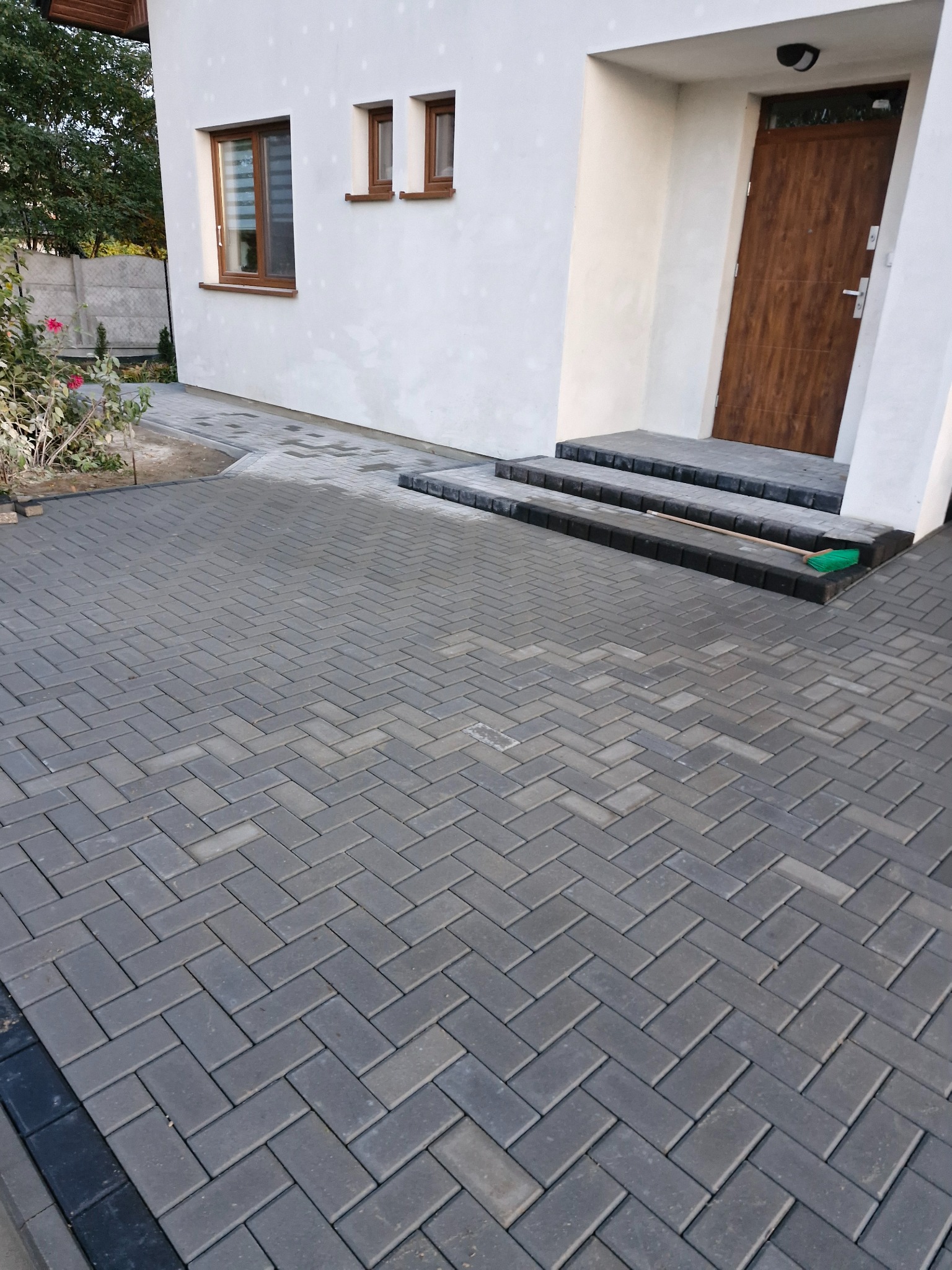 Nowo ułożona szara kostka brukowa w jodełkę przed domem z drewnianymi oknami i drzwiami, z zieloną zmiotką opartą o schody.