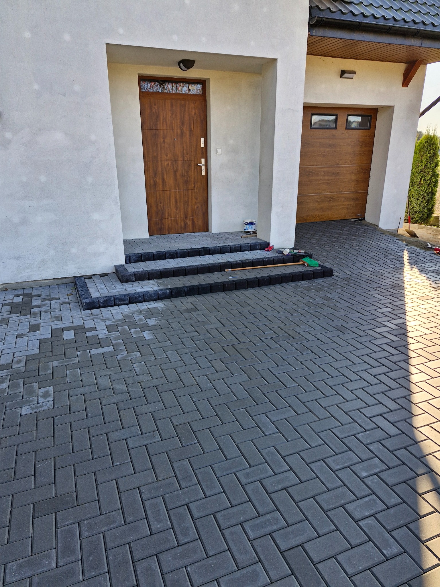 Ułożona szara kostka brukowa w jodełkę prowadząca do wejścia domu z trzema schodami wykończonymi ciemniejszą kostką, widoczne narzędzia brukarskie.