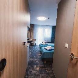 Wnętrze pokoju hotelowego z dwoma pojedynczymi łóżkami, biurkiem przy oknie i szafą w zabudowie, widoczne z perspektywy wąskiego korytarza z dekoracyjną podłogą.