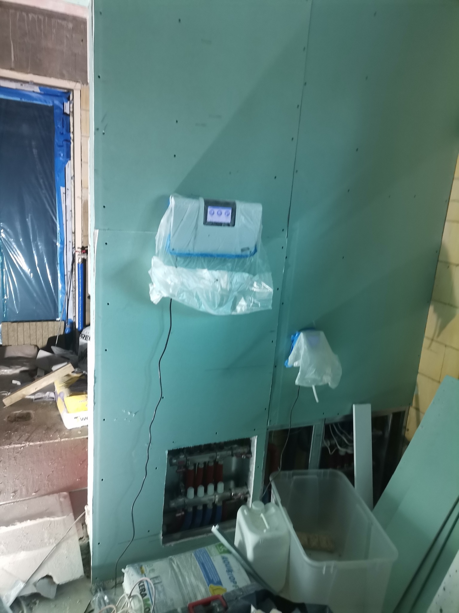 Montaż zielonej płyty karton gips na ścianie, widoczne otwory na instalacje, zabezpieczony folią sterownik ogrzewania podłogowego, narzędzia i materiały budowlane w nieładzie.