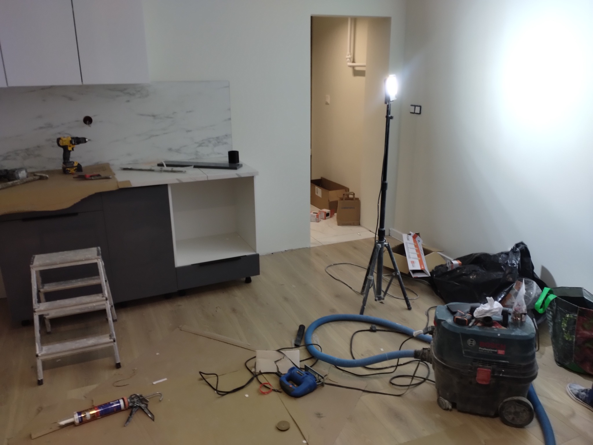 Wnętrze w trakcie prac wykończeniowych: zamontowany blat kuchenny z marmurowym wzorem, drabina, narzędzia, odkurzacz przemysłowy i lampa budowlana.