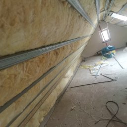 Artluk - Montaż izolacji z wełny mineralnej w metalowym stelażu na poddaszu, widoczne narzędzia i okno w tle.