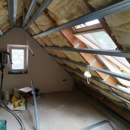 Artluk - Poddasze w trakcie adaptacji: widoczne ocieplenie wełną mineralną, metalowy stelaż pod zabudowę, okno dachowe z maską ochronną, przewody elektryczne i narzędzia na podłodze.