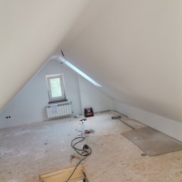 Artluk - Poddasze w trakcie prac wykończeniowych: białe ściany i skośny sufit, okno dachowe, odkryta klapa schodów strychowych, widoczne narzędzia i materiały budowlane na podłodze.