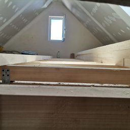 Artluk - Poddasze w trakcie adaptacji, widok z dołu na drewnianą konstrukcję stropu i częściowo wykończone ściany gipsowo-kartonowe z widocznymi łączeniami szpachlowanymi, małe okno dachowe w tle.