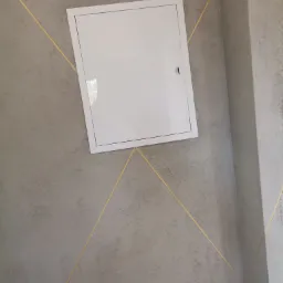 Szara ściana z efektem betonu, zdobiona geometrycznym wzorem z cienkich, złotych pasków, z wbudowaną białą klapą rewizyjną.