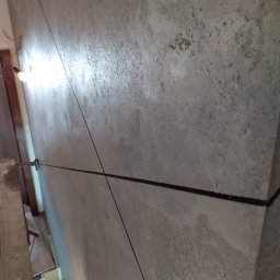 Artluk - Ściana z surowym wykończeniem, widoczne ślady szpachlowania i linie wyznaczające przyszły podział, oświetlona żarówką na kablu.