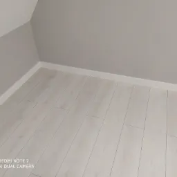 Jasne panele podłogowe ułożone w rogu pokoju z szarą ścianą, widoczne białe listwy przypodłogowe.